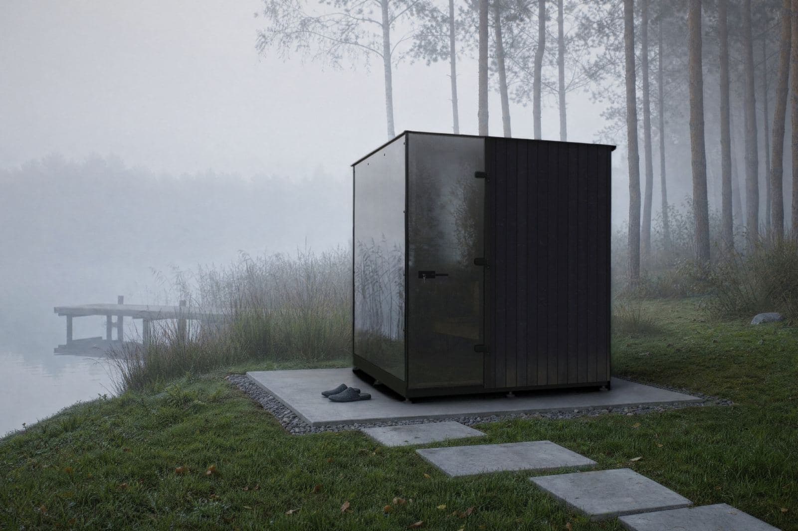 Sauna cubo con vetro fumé e rivestimento scuro su una lastra di pietra sulla riva nebbiosa di un lago, un pontile oltre le canne