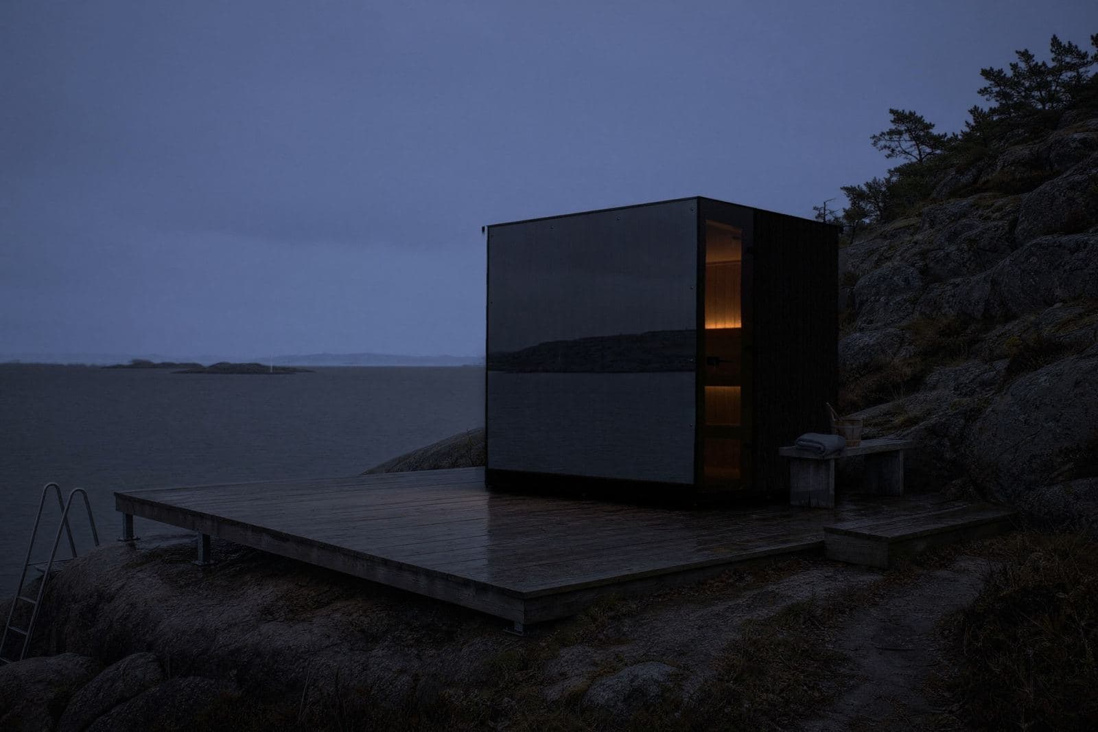 Sauna cubo con vetro a specchio su una terrazza in legno sopra una costa rocciosa al crepuscolo, luce calda all'interno