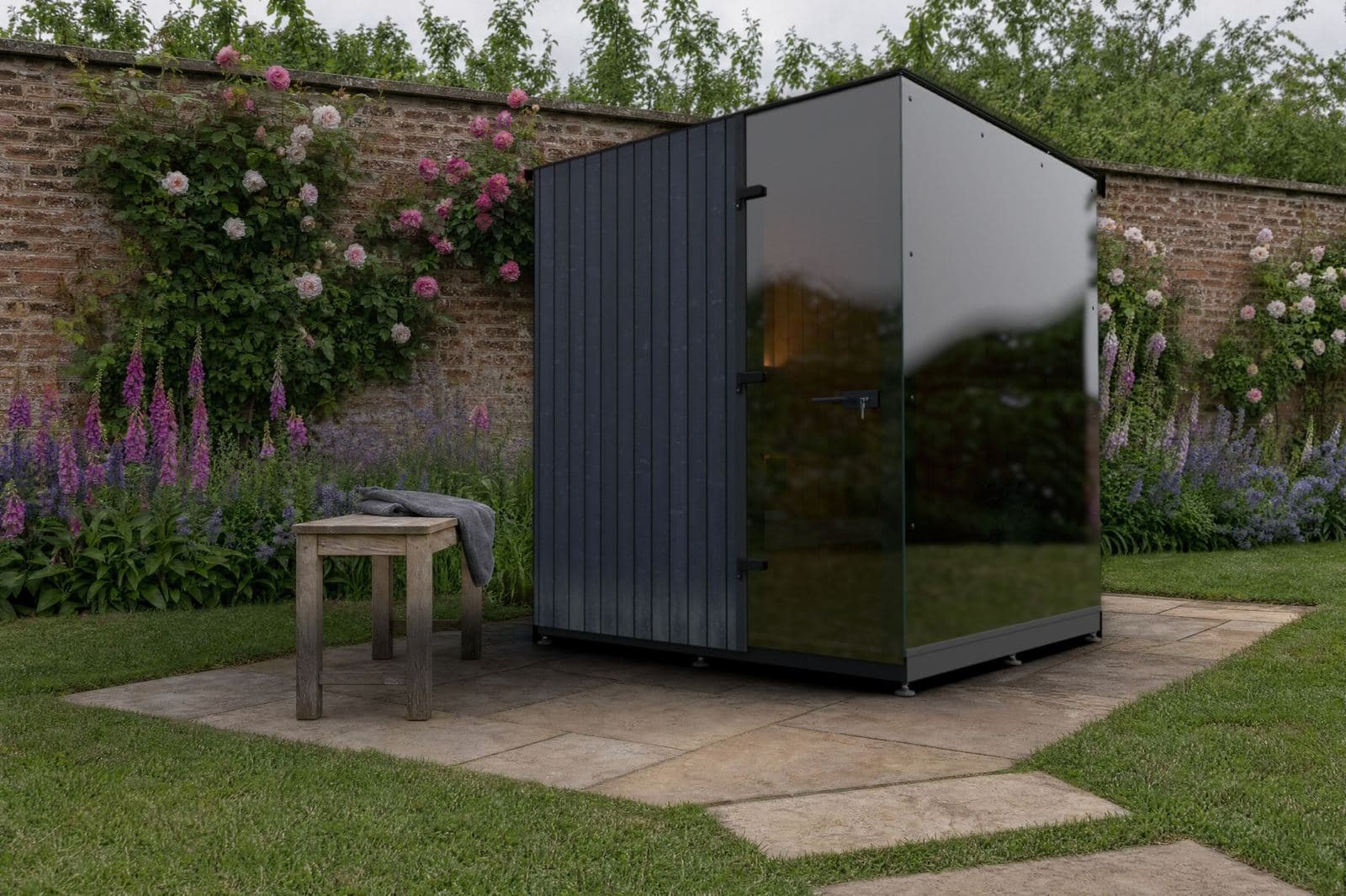 Sauna cubo con vetro a specchio su una terrazza in pietra in un giardino cinto da mura, con rose rampicanti, digitali e un tavolo in legno con un asciugamano